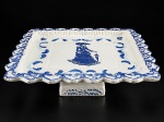 WEISS, um (1) prato para bolo de pé confeccionado em faiança vitrificada na tonalidade branca, decorados por elementos vegetalistas e dama antiga ao centro na tonalidade azul, bordas em ondulações, apresentam bicados e falhas na vitrificação, medindo 9,5x32x32cm.
