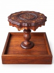 MOBILIÁRIO ORIENTAL, uma (1) mesa auxiliar de inspiração indiana, confeccionado em madeira entalhada, apresentando tampo circular com bordas recortadas e onduladas, ricamente ornamentado em marchetaria com motivos florais, arabescos e desenhos geométricos em composição concêntrica, com detalhes em tonalidades contrastantes. Estrutura sustentada por pedestal central torneado, com elementos arredondados, em acabamento castanho-avermelhado. Base quadrangular em madeira, com bordas elevadas e acabamento natural, medindo 40x38cm e base 47,5x47,5cm.