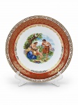 PORCELANA DE BAVARIA, um (1) prato fundo decorativo confeccionado em porcelana alemã na tonalidade branca, decorado por cena idílica em policromia, espessa faixa na tonalidade vermelha com guirlanda floral em vasta douração, leves falhas na douração, medindo 23cm de diâmetro. Pequeno fio de cabelo na borda.