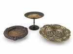 BRONZE e METAL DOURADO, lote com três (3) peças, sendo: A) 1 cinzeiro confeccionado em bronze com cinzelado de elementos vegetalistas, apoiada sobre 3 pés afilados, medindo 12,5cm, B) 1 cinzeiro com rico cinzelado de elementos vegetalistas e florais, medindo 16cm de diâmetro e C) 1 estrutura para castiçal confeccionado em metal dourado com ausência da bobeche e sinais de solda, medindo 8,5x11,5cm.