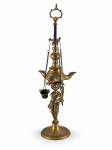 BRONZE, uma (1) lamparina confeccionada em bronze, com base circular, haste central ornamentada por figura humana em relevo, reservatório com dois bicos laterais e sistema suspenso por correntes, apresenta 3 utensilios utilizados no seu manuseio: extinguidor de lume, pinça para ativar o pavio e peça para óleo, medindo 39cm de altura.