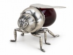 METAL PRATEADO e VIDRO, um (1) porta-mel italiano no formato de abelha com abdômen em vidro vermelho e montagem em metal espessurado a prata. Tampa com dobradiça, no formato das asas e apoio sobre três pares de pernas. Sob a base, numeração 16 com iniciais I.F.S. do artista e designer Israel Freeman & Son, de Milão. Início do séc. XX., apresentando pequeno bicado no recipiente, vide foto, medindo 9x15x9,5cm.