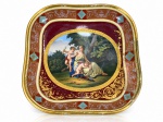 Royal Vienna (Áustria), um (1) centro de mesa quadrangular confeccionado em porcelana austríaca na tonalidade branca, centro pintura policromada com cena mitológica de Vênus e Amor (Cupido), em paisagem bucólica. Aba ornamentada em tons de vinho e terracota, com arabescos florais dourados, detalhes geométricos em azul-turquesa e bordas realçadas a ouro, apresentando falhas na douração e leves marcas do tempo, medindo 24x24cm.