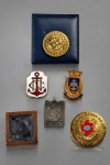 MILITARIA E MEDALHAS, conjunto de seis (6) peças entre distintivos, insígnias, medalhas e placas comemorativas, confeccionadas em metais de diferentes tonalidades, com acabamentos dourados, prateados e detalhes em esmalte policromado, sendo: A) distintivo circular dourado com representação de roda dentada, verso ROTATORY NO BRASIL, acondicionado em estojo azul-marinho; B) distintivo esmaltado em branco, com âncora vermelha e monograma central dourado, alusivo à tradição naval; C) distintivo do COLÉGIO NAVAL, encimado por coroa e apresentando brasão esmaltado em azul, com inscrição institucional; D) placa quadrangular com representação de nu feminino em relevo, sobre base de tonalidade acobreada; E) medalha retangular em metal prateado, com representação de aeronave em relevo e ornamentações geométricas; F) medalha circular dourada, com brasão esmaltado em policromia e inscrições alusivas ao Estado do Rio de Janeiro. Conjunto de interesse histórico, militar, institucional e colecionável, apresentando marcas do tempo e de uso. Peças no estado, maior peça medindo 4x4cm e menor 3,2x2,5cm.