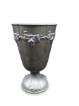 Vaso em Metal Ornamental com Guirlanda de Uvas de corpo gomado. Apresenta faixa superior decorada com guirlandas em relevo com motivos florais e cachos de uvas, além de ornamentação combinada na base circular.  Medidas: 22 cm (altura) x 13 cm (diâmetro) Apresenta pátina natural do tempo, marcas de oxidação e desgastes característicos do uso.