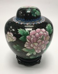 China / Beijing Cloisonné. Imponente potiche decorativo em preto, elaborado em autêntico Beijing Cloisonné, técnica tradicionalmente associada ao luxo imperial. A peça possui alto valor artístico e histórico. Sobre o corpo de cobre, são soldados pequenos fios que formam os compartimentos (cloisons), posteriormente preenchidos com pigmentos de esmalte para compor o design. O pote e a tampa passam por múltiplas etapas de queima e polimento até atingirem o acabamento final. A decoração é detalhada, apresentando flores de peônia em rosa e branco, peônias em azul e branco, além de flores de cerejeira no verso e folhagens verdes. As tiras de cobre que formam os compartimentos são visíveis, conferindo um relevo característico à peça. Acompanha base de madeira ricamente entalhada. Período: Século XX. Marca: "Made in Beijing, China" na base. A borda na base do potiche apresenta pequenos desgastes naturais na douração, compatíveis com a idade da peça. Dimensões: Altura do potiche: 18 cm | Altura total (com base): 22 cm.