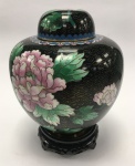 China / Beijing Cloisonné. Imponente potiche decorativo em preto, elaborado em autêntico Beijing Cloisonné, técnica tradicionalmente associada ao luxo imperial. A peça possui alto valor artístico e histórico. Sobre o corpo de cobre, são soldados pequenos fios que formam os compartimentos (cloisons), posteriormente preenchidos com pigmentos de esmalte para compor o design. O pote e a tampa passam por múltiplas etapas de queima e polimento até atingirem o acabamento final. A decoração é detalhada, apresentando flores de peônia em rosa e branco, peônias em azul e branco, além de flores de cerejeira no verso e folhagens verdes. As tiras de cobre que formam os compartimentos são visíveis, conferindo um relevo característico à peça. Acompanha base de madeira ricamente entalhada. Período: Século XX. Marca: "Made in Beijing, China"  Estado de conservação: Excelente. Dimensões: Altura do potiche: 18 cm | Altura total (com base): 22 cm.