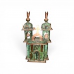 Miniatura de Igreja Colonial decorativa em cobre e latão, representando fachada de igreja barroca colonial. Apresenta duas torres sineiras vazadas com sinos móveis, cúpulas bulbosas com pináculos e frontão central encimado por cruz latina. Belíssimo acabamento com pátina esverdeada (azinhavre) em contraste com o metal polido. Provável manufatura mineira ou de arte sacra popular. Medidas: 22 x 14 cm.