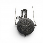 Antigo polvirilho Heráldico  em metal fundido e patinado (formato "bolacha"), ricamente decorado em relevo sobre fundo granulado. Face A: Brasão coroado com três flores-de-lis, ladeado por águias como suportes e volutas barrocas. Face B: Elmo de cavaleiro com ricos paquifes e escudo inclinado com águia imperial. Apresenta gargalo com alça estilizada e passadores laterais. Europa, estilo Historicista (séc. XIX). Medidas: 23 x 18 cm , marcas do tempo e pátina natural do metal. Fios de arame substituindo as argolas originais de suspensão.