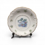 Prato Decorativo em Porcelana Alemã KPM borda recortada com relevos rococó em motivos de rocaille e acabamento em filete de ouro. decorada com delicados ramos florais policromados. No centro, refinada cena em estilo Chinoiserie retratando paisagem oriental com ponte, pavilhão e vegetação. Alemanha, séc. XX. Excelente estado