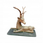 Escultura de cervo reclinado em bronze fundido e cinzelado  com expressiva estilização na galhada e na textura da pelagem do pescoço. Apresenta marcas incisas ornamentais nas laterais e pátina natural do tempo. Fixada sobre base de vidro retangular. Medidas: 44 x 42 cm e Base: 20 x 45 cm.