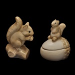 Conjunto decorativo temático de esquilos, composto por uma estatueta e um pote utilitário em cerâmica esmaltada, sendo A) Estatueta decorativa de esquilo posicionado sobre um pedaço de tronco de árvore. O animal é retratado segurando uma pequena noz ou semente entre as patas dianteiras. 17 x 12 x 7 cm. B) Pote  no formato de noz, com tampa removível. No topo da tampa, há um pequeno esquilo sentado servindo como puxador da peça. 16 x 13 x 10 cm. No verso marca da manufatura nacional Luiz Salvador, Itaipava, RJ.