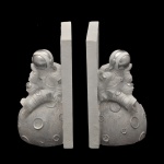 Par de serre livre em madeira pintada, temáticos de astronauta, projetados para manter livros organizados verticalmente em prateleiras ou mesas de forma criativa. Cada peça apresenta uma figura detalhada de um astronauta sentado sobre uma superfície que imita a textura e os relevos da Lua. Arte decorativa e de design moderno.