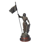 Escultura em petit bronze e outas ligas retratando a figura de  Joana d'Arc com armadura, escudo e estandarte. Retrata a icônica heroína e santa francesa em uma pose triunfante, segurando seu estandarte militar e apoiando a mão esquerda sobre um escudo decorado com motivos heráldicos. Base circular. Apresenta um dos pés quebrado. 47 x 16 x 15 cm.