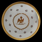 SÈVRES  Prato em porcelana francesa, homenageando o Imperador Napoléon 1º. Apresenta borda dourada e margem larga ornada por adornos florais repetidos. Centro ornamentado por águia coroada, emblema característico do imperador, emoldurada por coroa de louros. Sob a base, a inscrição à mão Manufacture Impériale de Sèvres e pequena coroa dourada encimada por cruz. 30 cm.