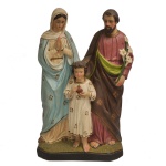 FÁBRICA BOM PASTOR - Escultura sacra em estuque (revestimento feito à base de gesso, areia e pó de mármore), policromada e dourada representando a Sagrada Família com o Menino Jesus ao centro da cena, segurando o Sagrado Coração, símbolo do amor divino e misericordioso de Deus para os homens desde a infância. N. Sra é retratada com as mãos reunidas em oração, vestindo o seu tradicional manto azul celeste. Do lado oposto, São José com os lírios brancos que refletem a pureza e a castidade do santo, com sua missão de ser o protetor da família. Base retangular chanfrada. Marca da manufatura sob a base. 41 x 23 x 11 cm.