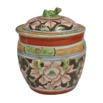 Pote com tampa em cerâmica chinesa policromada e esmaltada com intensa decoração de peônias. Pega no topo da tampa tem o formato esculpido de um cão-leão budista (quilin) acompanhado por pequenas alças laterais em formato de máscara. As famílias Peranakan consideravam os kamchengs itens valiosos de prestígio, utilizados principalmente em casamentos e ocasiões festivas. 22 x 21 cm.