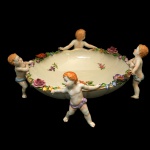 Centro de mesa em porcelana alemã policromada, de formato circular fundo, sustentada por figuras de meninos. Interior ornamentado por buquês florais. Borda realçada por flores e folhagens em alto relevo. Sob a base, marca da manufatura von Schierholz  Handmalerei. 13 x 32 cm.