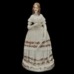 Escultura em porcelana policromada alemã, retratando uma dama da corte com vestido de época. Exibe belo vestido rendado e bordado com faixas floridas, segurando um carnet de bal na mão. Base circular. Marca da manufatura no verso. 43 x 22 x 22 cm.