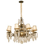 Lustre francês em bronze dourado e cristal. A peça apresenta uma estrutura robusta com 8 braços recurvados em bronze que sustentam bocais para lâmpadas, cada um decorado com pequenas cúpulas (mangas) de tecido bege claro. O design é ricamente adornado por pingentes, prismas e placas de cristal lapidado multifacetados, terminando em uma esfera de cristal lapidado na base central. Acompanham mini cúpulas cônicas em tecido claro com acabamento sutil nas bordas. 110 x 70 cm.