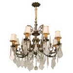 Lustre em cristal e bronze no estilo francês, com estrutura clássica ornamentada e acabamento envelhecido. O modelo conta com 8 braços curvilíneos que sustentam cúpulas de tecido e uma rica cascata de pingentes e placas de cristal lapidado. Haste robusta em liga metálica fundida (bronze) com detalhes florais e volutas. A corrente superior é revestida por uma capa de cordão decorativa bege, ocultando a fiação elétrica. Pequenas mangas em tecido claro de formato cônico, que suavizam a iluminação direta das lâmpadas. 120 x 72 cm.