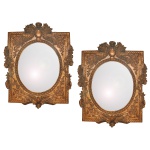 Belíssimo par de espelhos com moldura em madeira entalhada e dourada, apresentando um formato oval com acabamento bisotado. A peça exibe características marcantes do estilo clássico europeu, provavelmente de origem francesa, combinando um painel central oval cercado por uma moldura retangular adornada com elementos florais, ramos e arabescos em relevo. 100 x 80 cm (MT). 58 x 47 cm (MI).