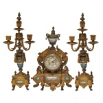 Garniture de cheminée em bronze ormolu, ornada por adornos em porcelana. Constando de um relógio circular com mostrador branco, ladeado por águias de asas abertas e encimado por pequeno vaso com tampa pontuda. Base construída com segmentos laterais recortados e recamados por folhagens com elemento retangular ao centro, ornamentados por placas policromadas decoradas por paisagens rurais com figuras. Frente realçada por guirlanda com flores. Pés curtos, revirados em garras. Acompanham par de candelabros para 3 velas, montados com adornos cônicos e alongados, bordados por colares floridos. Hastes retas com anelados, sustentando braços recurvos rematados por bobeches com suportes salientes, centralizando pequenos castiçais com ponteiras nos topos. Bases quadrangulares, acompanhando a decoração, com pequenos painéis policromados. Quinas encimadas por pinhas e ornadas por colunatas sobre pés recurvos. 40 x 28 cm (relógio). 47 x 18 cm (candelabros).