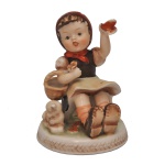Estatueta em porcelana europeia, retrata uma menina sentada acenando com um lenço vermelho, acompanhada por um pequeno cordeiro e uma cesta. Pintura à mão com aspecto fosco. Sem marca. 12 x 10 x 10 cm. Espólio de Josephina Penteado da Fonseca, SP.
