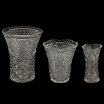 Lote constando de trio de vasos em cristal lapidado à mão em diferentes tamanhos e ricos padrões geométrico. A)  Apresenta uma lapidação clássica e profunda em formato de bico de jaca (losangos em relevo) cobrindo todo o seu corpo cônico, terminando em uma borda reta facetada. Pequeno bicado na borda. 20 x 15 cm. B) Possui formato com silhueta levemente facetada e borda ondulada recortada, decorado com um rico trabalho que mistura sulcos verticais, rosetas e estrelas.16 x 14 cm. C) Um delicado vaso de flores estilo bud vase com borda recortada em arcos e lapidação vertical acentuada, ideal para pequenos arranjos de mesa. 15 x 8 cm. Espólio de Josephina Penteado da Fonseca, SP.
