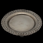 Salva circular em metal prateado, caracterizada centro levemente rebaixado com aba larga voltada para fora. Borda cinzelada com detalhes em relevo no formato de conchas. 28 cm. Espólio de Josephina Penteado da Fonseca, SP.