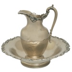 Conjunto de gomil em metal prateado composto por uma jarra com alça curva com apoio para os dedos e uma bacia redonda. Apresentam borda decorada com pequenas rosas e arabescos. Marcas de uso e pátina do tempo. 13 x 46 cm (jarra). 26 x 37 cm. Espólio de Josephina Penteado da Fonseca, SP.