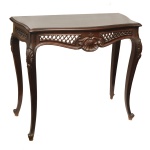 Mesa de encostar em madeira nobre, lavrada em estilo neorrococó. Tampo retangular com laterais e frente onduladas. Saia esculpida e vazada em treliças, centralizando adorno concheado à frente. Pernas curvilíneas, com joelhos salientes realçados por folhagens e rematados por pés revirados sobre sapatas. Manchas de uso. 90 x 100 x 44 cm. Espólio de Josephina Penteado da Fonseca, SP.