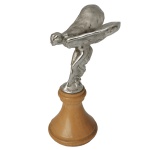 Miniatura de capô Spirit of Ecstasy, conhecida como "Emily" ou "The Flying Lady", símbolo clássico da Rolls-Royce, originalmente esculpida por Charles Sykes. A peça metálica prateada está fixada sobre uma base de madeira torneada. 23 x 12 x 9 cm.