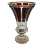 Vaso em cristal da Boêmia, moldado no formato de corneta facetada, ornado por faixas de cor rubi e realçado por faixa dourada. Base lapidada, com anelado largo e saliente, apoiado em haste curta sobre pé de mesmo estilo com borda recortada. 24 x 15 cm.