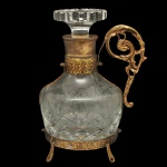 Licoreira em cristal lapidado com base e alça trabalhada em metal dourado. Apresenta um elegante design clássico com suporte de pezinhos.24 x 17 cm.