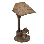 Luminária de mesa em ferro patinado, com design figurativo orientalista. Apresenta base na forma de  escultura detalhada retratando um camelo com sela e uma figura humana ao seu lado sob uma estrutura que imita o tronco e a cobertura de uma cabana ou palmeira estilizada. Fiação precisando de reparo. 25 x 12 x 12 cm.