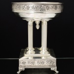 METAL PRATEADO, uma (1) imponente fruteira de pé alto, base e barrado da borda com ornamentação relevada em parras, prato circular assente sobre quatro colunas caneladas na vertical de capiteis lisos, base trapezoidal, pés em garra, detalhe central em bilro entre as colunas, sem marca de manufatura aparente, medindo 25 cm diâmetro e 32 cm altura, vide foto.