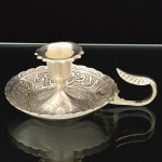 METAL PRATEADO, uma (1) palmatória com alça, ornamentação cinzelada no padrão Império Otomano com elementos geométricos e vegetalistas, além de legendas em árabe, medindo 10 cm diâmetro e 5 cm altura, vide foto.