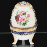 PORCELANA, uma (1) caixa no formato ovoide, ornamentação policromada com rosas, faixas na tonalidade azul cobalto, arremates a ouro com pérgulas, volutas e elementos vegetalistas, marca Fine Porcelain Hand Painted  24K Gold Deco Collections LIMOGES, monture em metal dourado, base trípode, medindo 15 cm altura, vide foto.