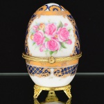 PORCELANA, uma (1) caixa no formato ovoide, ornamentação policromada com rosas, faixas na tonalidade azul cobalto, arremates a ouro com pérgulas, volutas e elementos vegetalistas, marca Fine Porcelain Hand Painted  24K Gold Deco Collections LIMOGES, monture em metal dourado, medindo 9,5 cm altura e 6 cm diâmetro, vide foto.