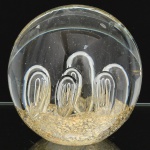 VIDRO, um (1) peso de papel quase esférico, incolor e translúcido, interior com bulicados ovoides e pó de ouro, confeccionado na técnica dos Grandes Mestres Vidreiros de Murano, medindo 10 cm altura, vide foto.