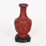 ARTE ORIENTAL, um (1) vaso no formato de balaustre, confeccionado em metal, recoberto em laca vermelha com entalhes representando dragões celestiais, nuvens e elementos vegetalistas, interior esmaltado na tonalidade azul, acompanha peanha em madeira torneada e ebanizada, altura total 17,5 cm, vide foto.