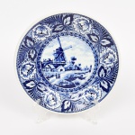 PORCELANA HOLANDESA, um (1) prato decorativo, ornado com paisagem representando marina, barcos, moinhos e figuras, borda com barrado em elementos vegetalistas, tonalidade azul e branca, marca By BOCH  DELFTS, medindo 23 cm diâmetro, vide foto.
