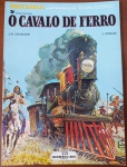 HQ Rara: Tenente Blueberry  O Cavalo de Ferro (Charlier / Giraud)  Ed. Meribérica/Liber"O Cavalo de Ferro" (Le Cheval de Fer) inicia um dos arcos mais aclamados e maduros de toda a série Blueberry: a epopeia da construção da linha ferroviária transcontinental americana. Na trama, o herói é designado para pacificar as tribos indígenas e evitar que os trilhos do progresso (batizados pelos nativos como "o cavalo de ferro") desencadeiem uma guerra sangrenta, enfrentando no caminho traições corporativas e sabotagens. A arte de Jean Giraud neste volume atinge níveis extraordinários de detalhes técnicos e de ambientação. Uma joia editorial da extinta editora Meribérica, totalmente fora de catálogo há décadas e muito cobiçada.