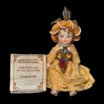 CREAZIONE MARIGIO - Boneca em biscuit e trajes detalhados de época. Destaca-se pelo vestido rendado amarelado e um adorno de cabeça volumoso com uma folha decorativa no topo. No peito, ela traz um ornamento com pedrarias simuladas em tons de âmbar e vermelho. Ao lado esquerdo, encontra-se o folheto do Certificado de Garantia, indicando sua autenticidade como objeto de coleção. 10 x 6 cm.