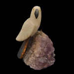 Escultura artesanal de um papagaio esculpida em quartzo branco com bico texturizado e apoiada sobre uma drusa de ametista natural. 10 x 7 x 7 cm.