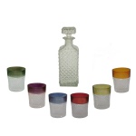 Conjunto para bar em vidro composto por garrafa licoreira e seis copos. A) garrafa de formato quadrado, em vidro com lapidação bico de jaca. Gargalo estreito e tampa geométrica lapidada. 24 x 8 x 8 cm. B) Conjunto de seis pequenos  copos de dose ou rocks, caracterizados por uma faixa colorida na borda superior. Cada copo apresenta uma tonalidade diferente na borda, incluindo tons de verde, roxo, azul, vermelho, amarelo e âmbar, contrastando com a base em vidro translúcido fosco. 9 x 8 cm Apresenta tampa quebrada por dentro.
