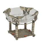 Centro de mesa em estilo eclético composto por uma bacia em vidro translucido integrada a uma base em metal. Bacia circular com bordas decoradas por adornos metálicos figurativos que parecem representar rostos ou elementos mitológicos estilizados nas extremidades. O suporte é sustentado por quatro colunas verticais com texturas aneladas, que repousam sobre plataforma quadrangular de metal trabalhado. Base inferior, pequenos detalhes em tons avermelhados dispostos ao longo do platô, conferindo uma estética única e sofisticada à peça. 8 x 11 cm.