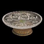 Fruteira estilo tazza em vidro prensado translucido e lapidado decorado com motivos geométricos e florais. A peça possui uma haste central com fixação metálica esverdeada e uma base de metal canelada com pátina envelhecida. 15 x 6 cm.