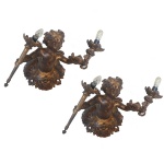 Par de apliques de parede para 2 luzes em bronze dourado, moldados no formato de bustos de putti, segurando tochas rematadas por bobeches e lâmpadas. Apresentam-se com os braços à frente, vestindo saiotes bordados. 28 x 27 x 23 cm.